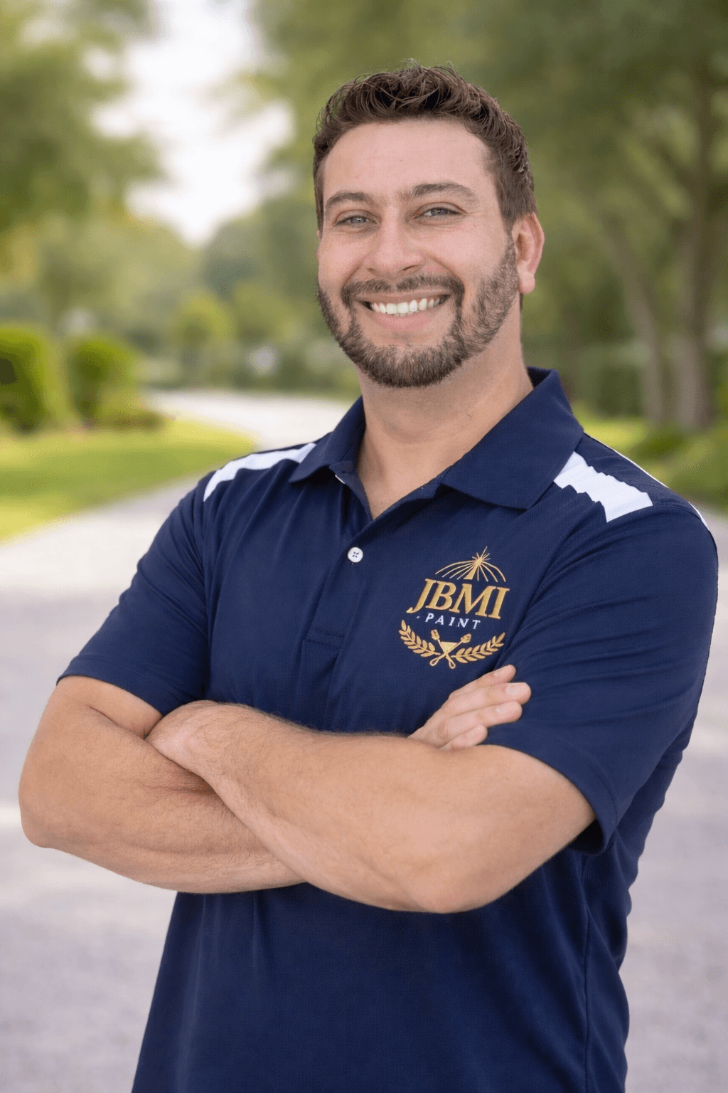 A bearded man in a navy JBMI Paint polo shirt smiles with arms crossed outdoors.